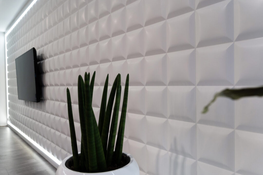 seitliche Sicht auf schlichte Pflanze in weißem Topf vor Wand aus weißen Kacheln, Fernseher im Hintergrund